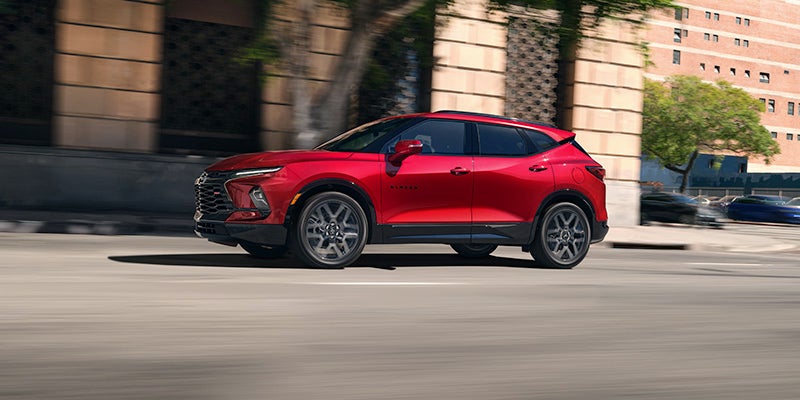 Red Chevrolet Blazer SUV driving along a city roadway, shown in side profile with modern buildings