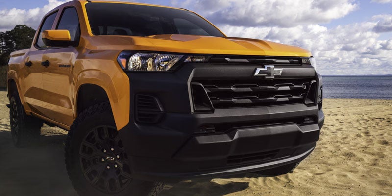 Orange Chevrolet pickup truck with a black grille on a sandy beach, ocean and sky in background.