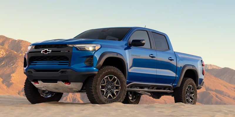 Blue Chevrolet pickup truck parked on a rocky desert landscape with mountains in the background, viewed from the front three-quarter angle