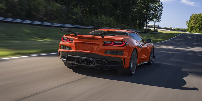 Rear three-quarter view of an orange Chevrolet Corvette sports car driving on a road bordered by trees and guardrails