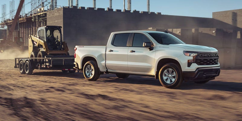 White Chevrolet Silverado pickup truck towing construction equipment on a trailer through a dusty worksite