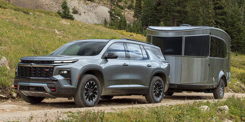 Gray Chevrolet Traverse SUV towing a silver travel trailer on a rugged mountain trail surrounded by grass